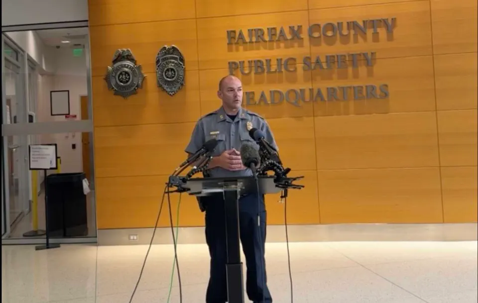 Police officer stands at a podium with multiple microphones in Fairfax County Public Safety Headquarters lobby.