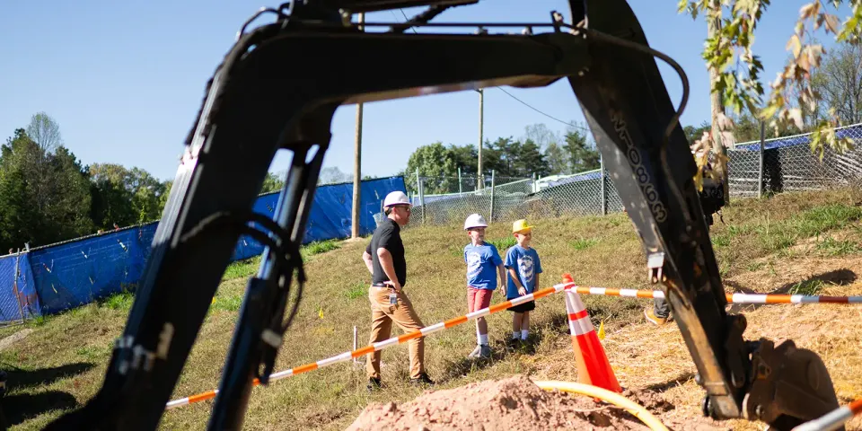 A dad shows his sons around a construction site, back hoe in the foreground