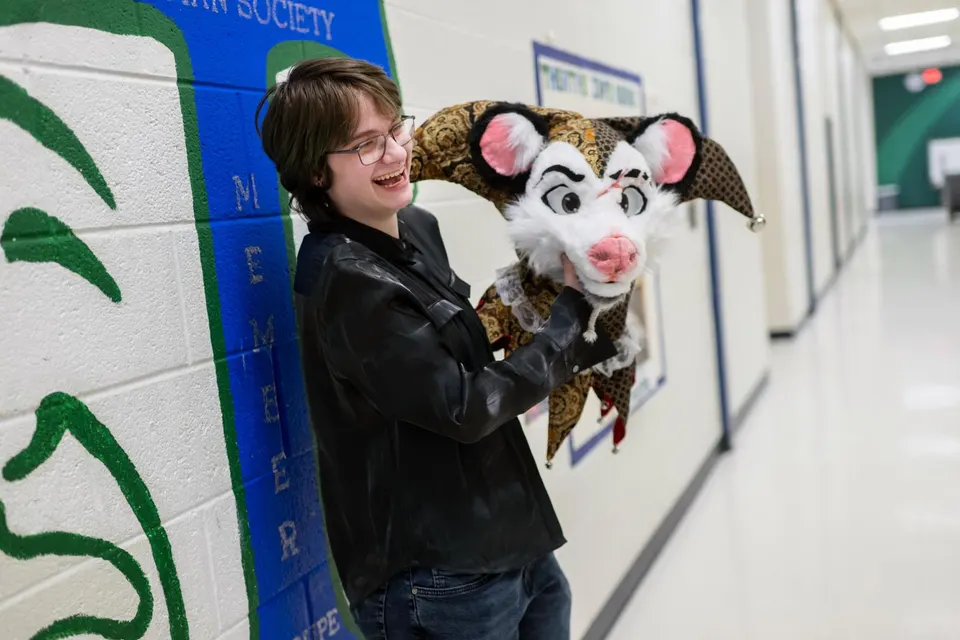 Dexter laughs in front of the Thespian Society mural in the hallway as he hold the head of a jester opossum he created.