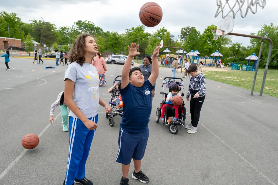 A boy looks up as he tosses a basketball in the air. A taller girl next to him looks on.