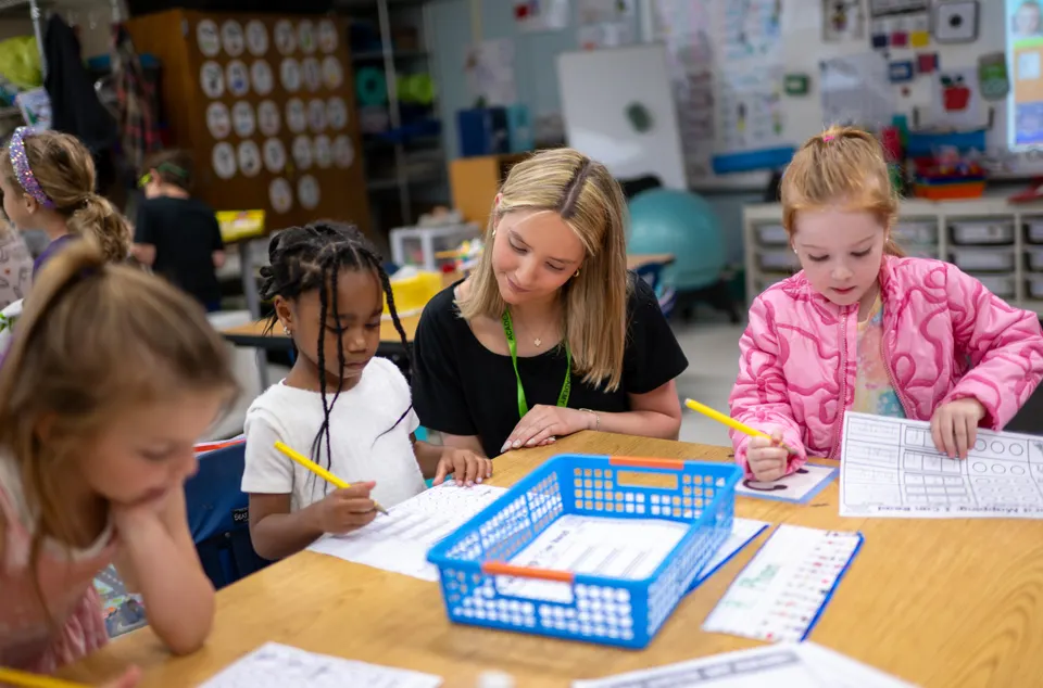 Alexa sits with three kindergarten students at their classroom table, helping them complete their literacy worksheets.
