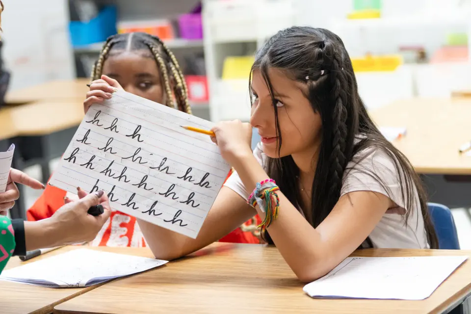 A student smiles as she points to a whiteboard covered in the cursive letter "h."