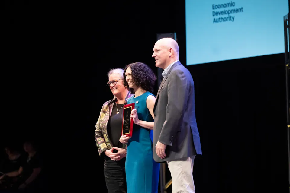 A woman (center) holds an award she just received from a woman and man on stage.