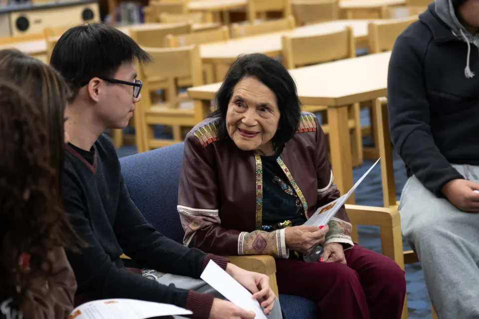 Dolores Huerta sits and smiles as she listens to a student speak.