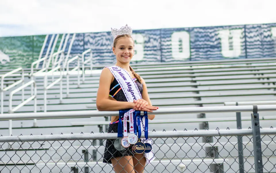 Mary stands in the bleachers, wearing a sash and crown. Several medals dangle on the fence in front of her.