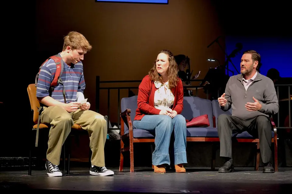 Three people sit on stage: a young man in a striped polo with backpack on the left, a woman in a red cardigan in the middle, and a man in a gray sweater speaking on the right.