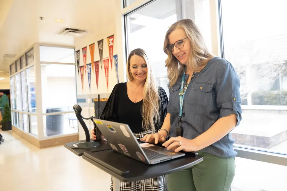 Two staff members review information together on a laptop in a school hallway.
