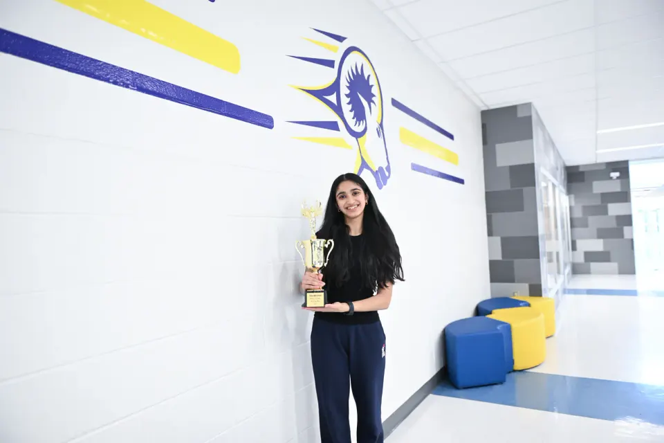 A student smiles, holding a trophy in a school hallway.