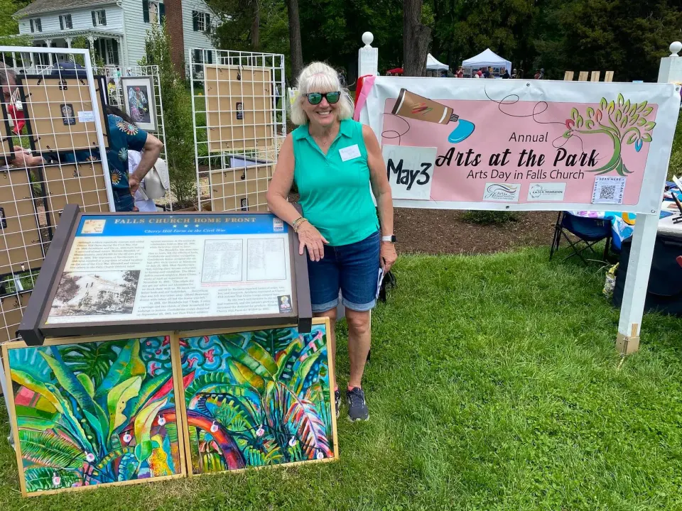 It's Arts Day in Falls Church! Roxanne Kaylor, principal organizer for Arts at the Park stands behind her own paintings (on grass) and before the banner she created for the...