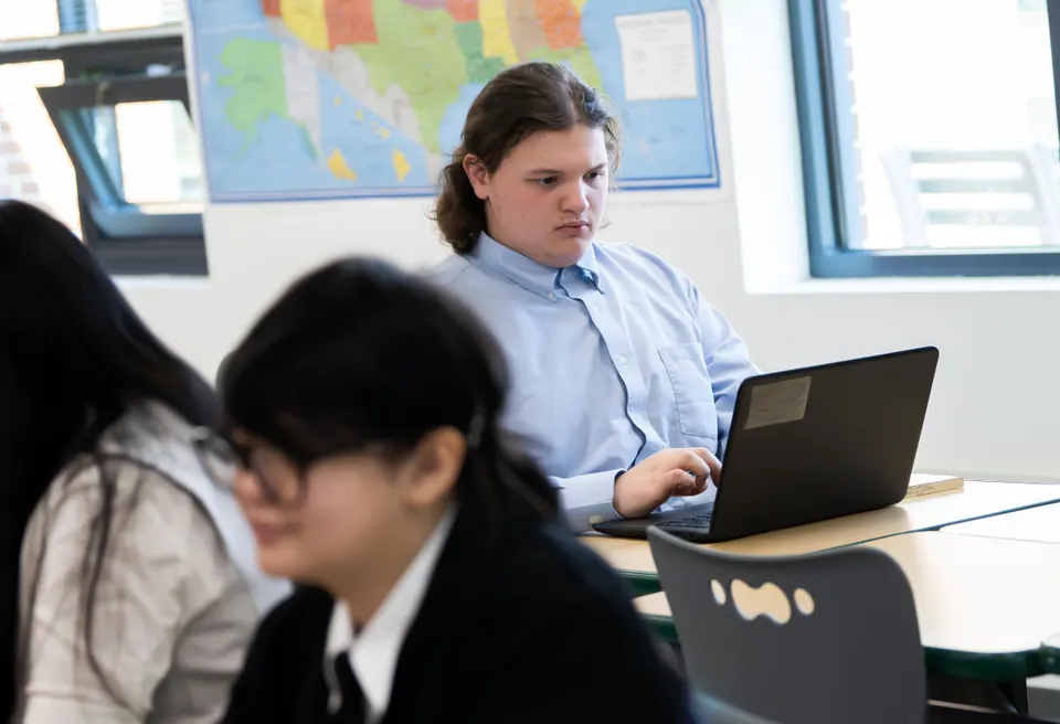 A high school student in a blue button-down shirt sits at a desk and types on a laptop.