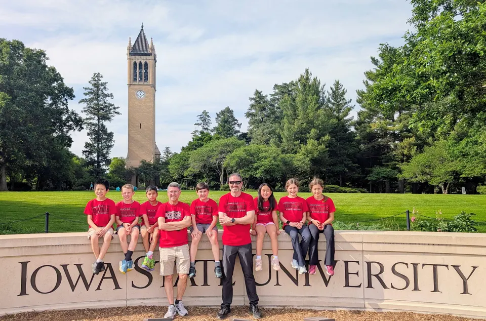 The students and their coaches pose for a photo sitting on, and standing in front of, the sign at the entrance to Iowa State University.