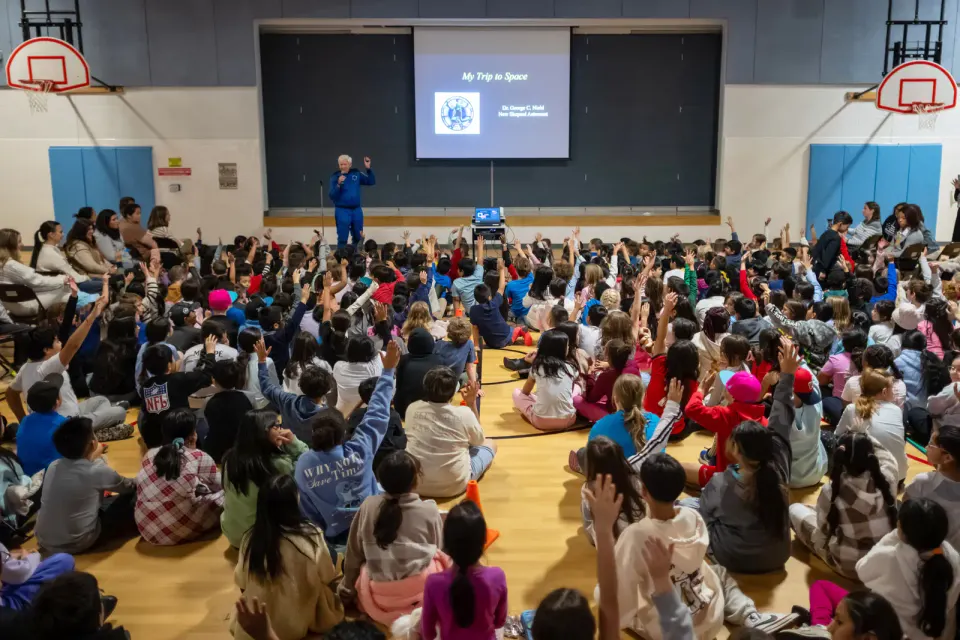 Hundreds of students raise their hands. They're looking at an astronaut in a blue jumpsuit, raising his left hand while holding a microphone in his right.
