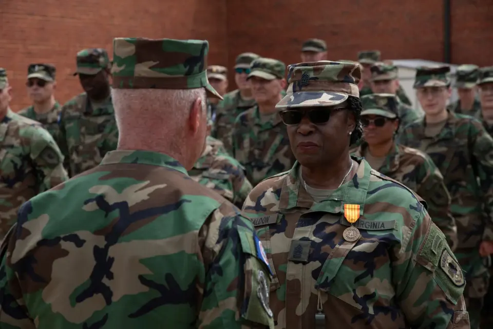 A woman in fatigues wearing a medal stands before a man in uniform