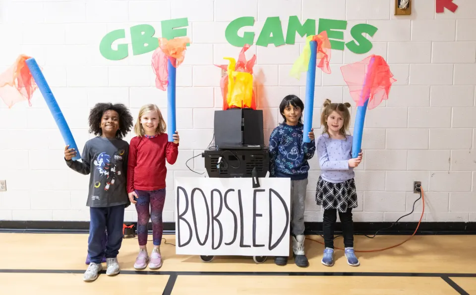 Students smile and hold "torches" made of pool noodles and flags. They stand on either side of a mock Olympic cauldron. Behind it, on the wall, green letters spell out: "GBE...