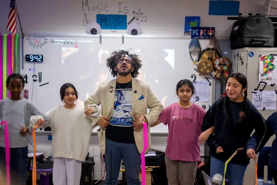 A teacher and students stand with arms linked, performing a call and response song