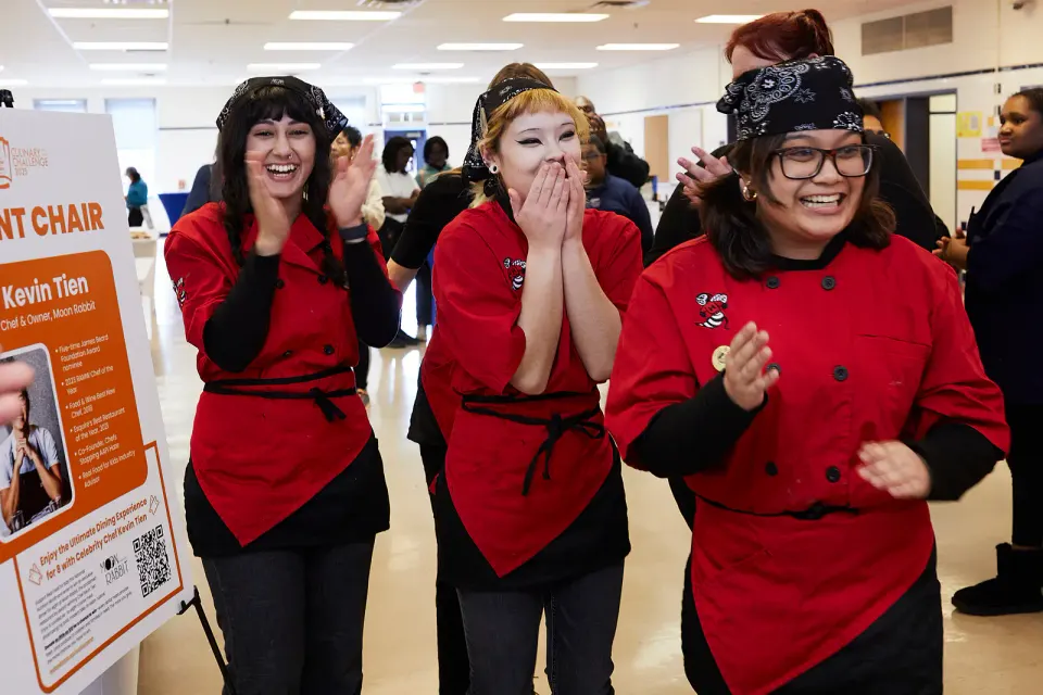Three students grin excitedly. One is clapping her hands, another has both hands held to her mouth, eyes wide open.