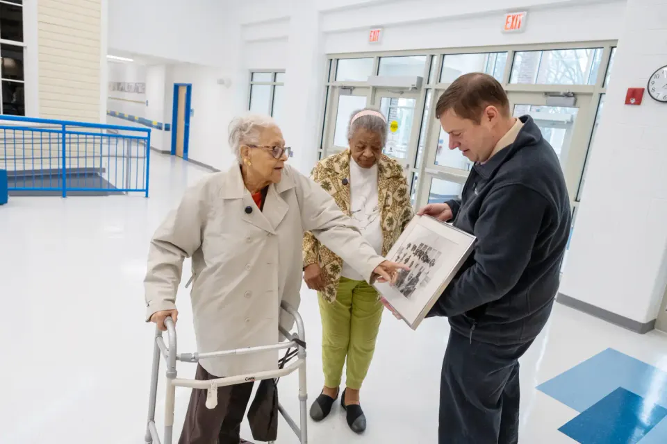A woman with a walker points to an old photograph in a frame held up by a man.