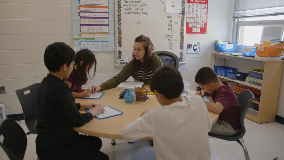 Teacher works with a small group of students at a table during a classroom reading and writing activity.
