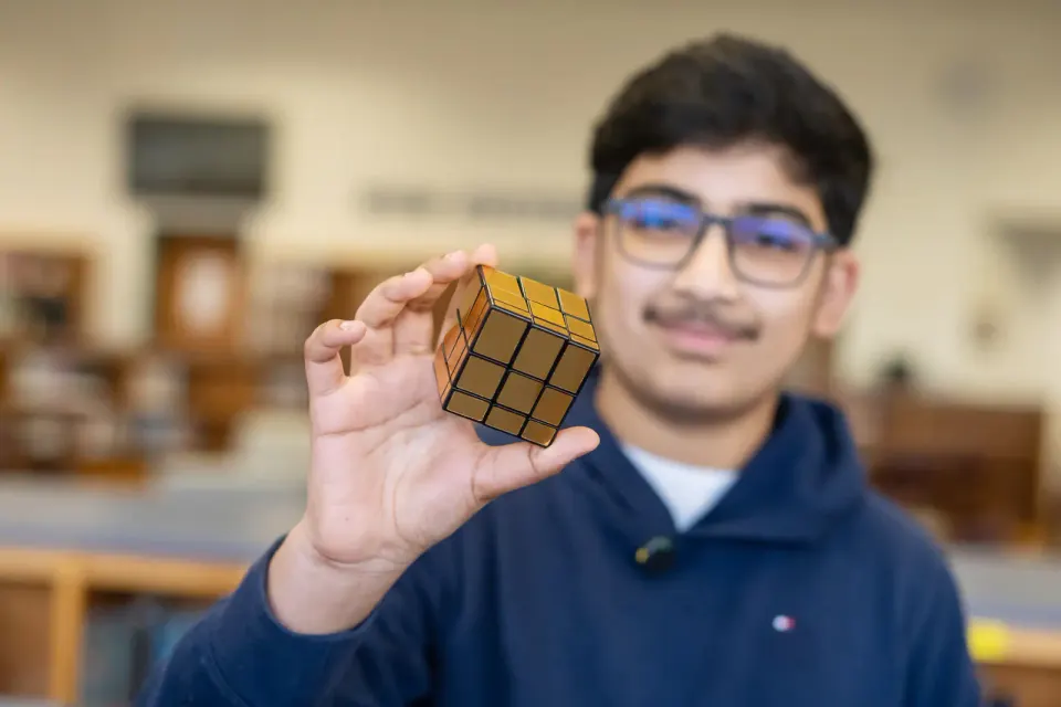 A student wearing glasses and a blue hoodie holds up a gold-colored puzzle cube.