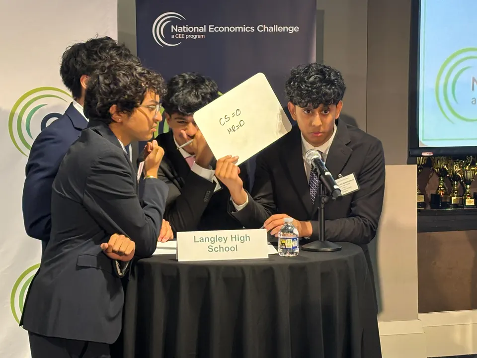 Four students in business suits huddle around a small table. One student holds up a small whiteboard while speaking into a microphone.