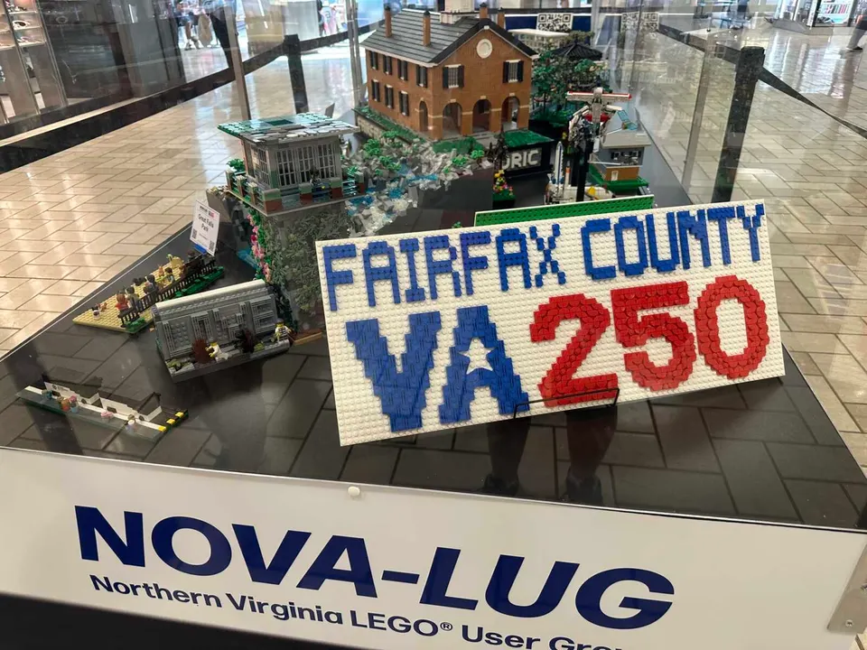LEGO display showing a town scene with a brick house and miniature structures, and a large sign reading 'FAIRFAX COUNTY VA 250' in blue and red bricks