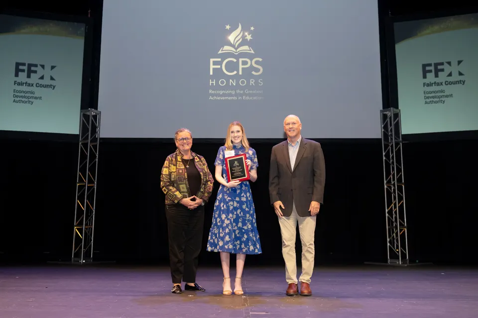 A teacher holding an award stands on stage between a man and a woman