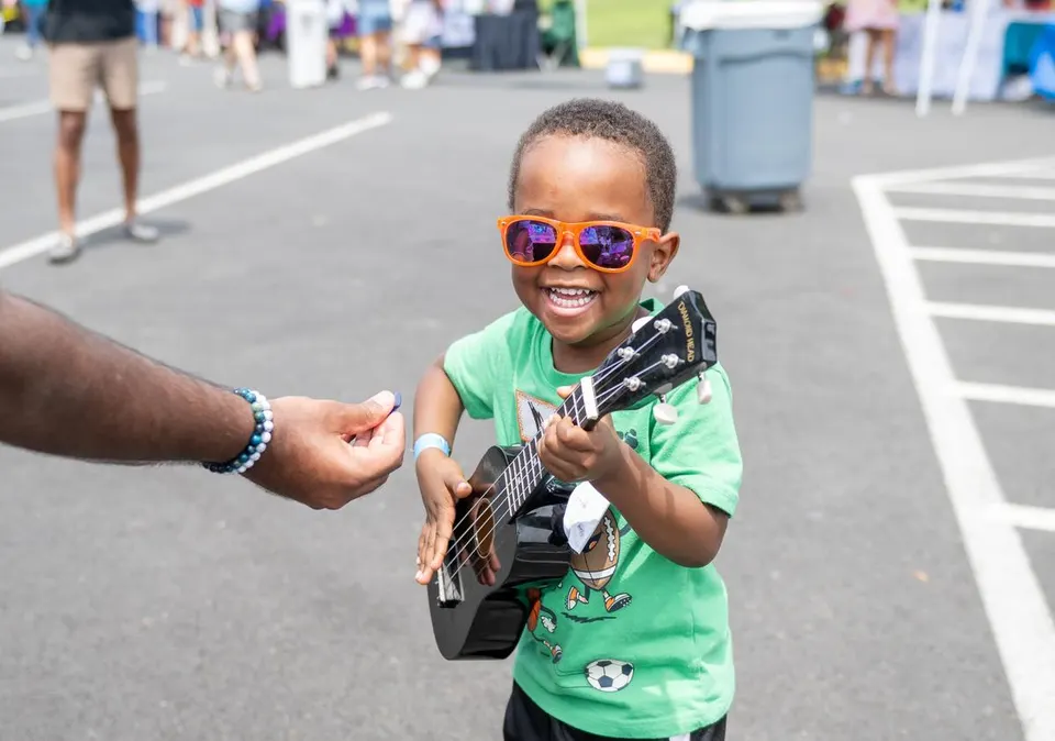 Child wearing sunglasses smiles while playing a small guitar at an outdoor event.