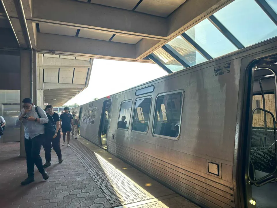 Silver metro train at the platform with doors open and passengers boarding under a concrete station canopy.