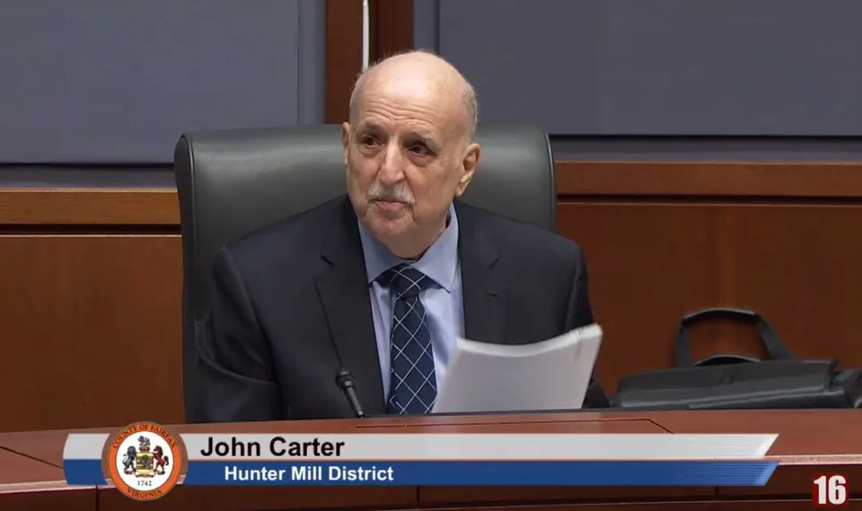 John Carter, Hunter Mill District official, sits at a wooden desk holding papers during a formal meeting, with a district seal visible on the left.