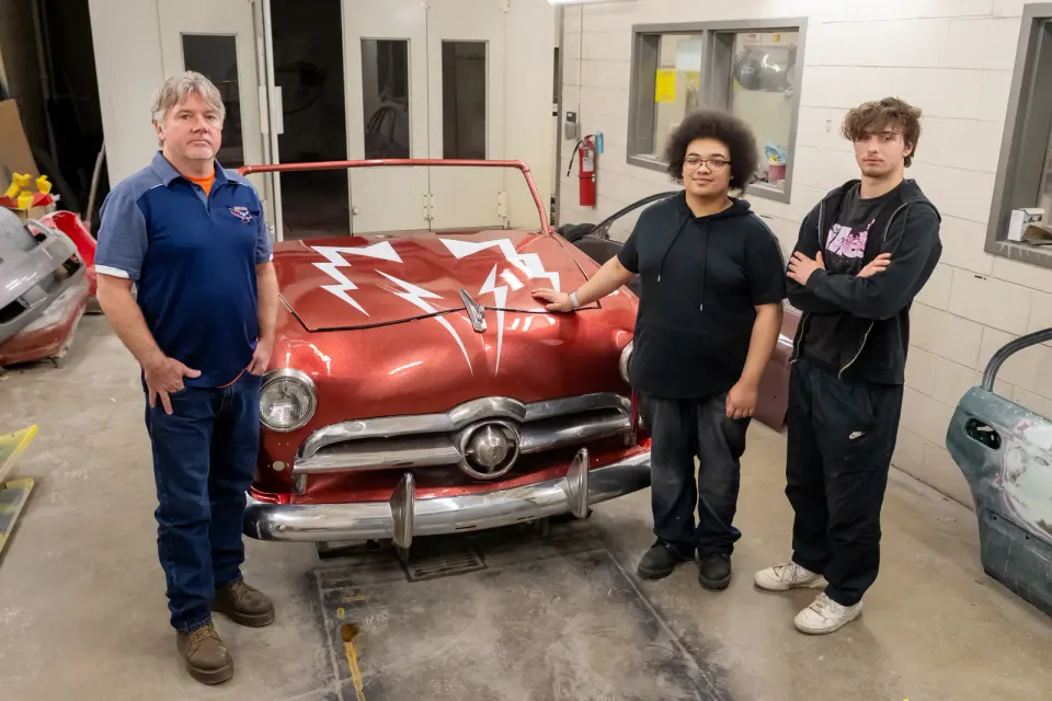 A man and two teenagers stand on either side of the front end of a red car.