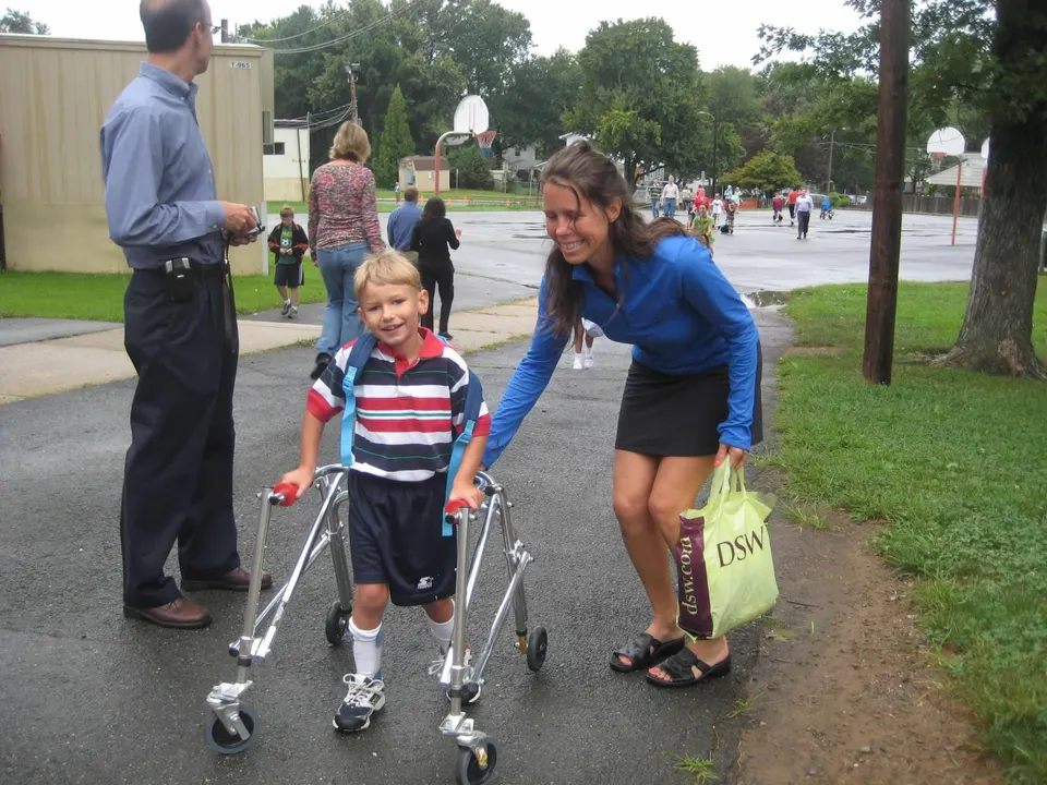 A mom stands behind a young boy using a walker who is wearing a backpack.