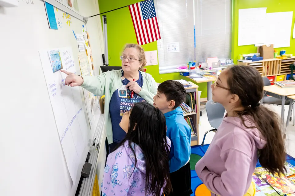 A teacher points at a picture taped to a whiteboard as three students look on.