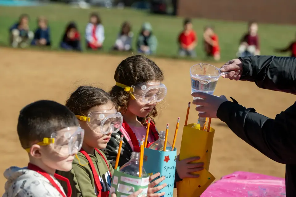Students wearing goggles hold bottles while vinegar is poured in