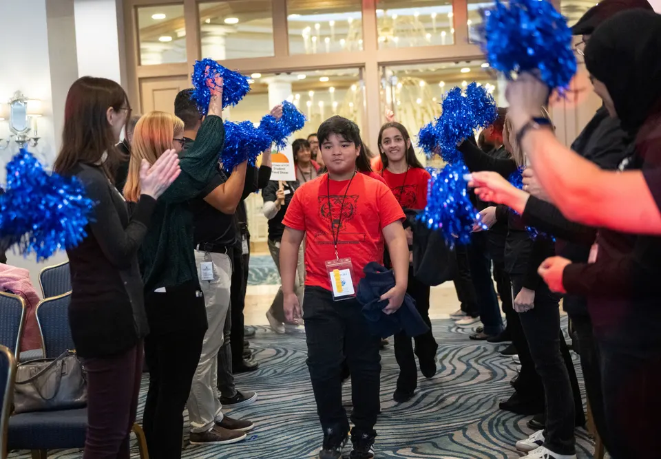 Students in red tshirts walk between two lines of people clapping and shaking pom poms.