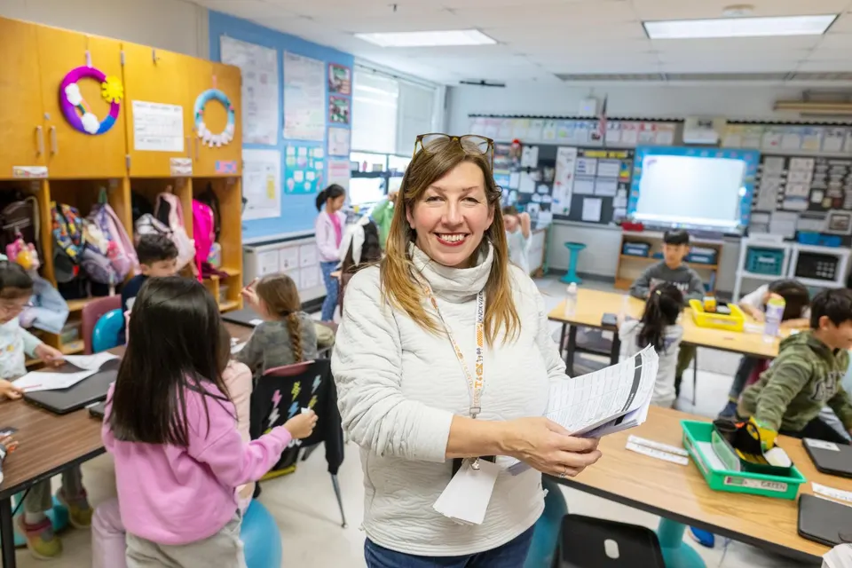 A teacher holding papers stands in front of a class, smiling.