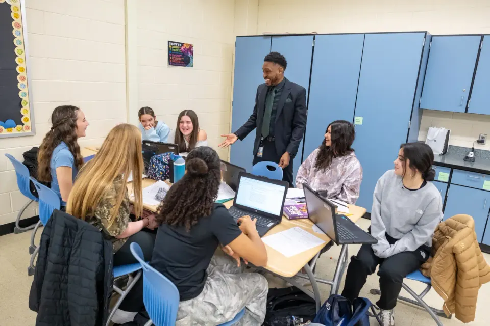 A man (standing) talks to a table of girls in a classroom