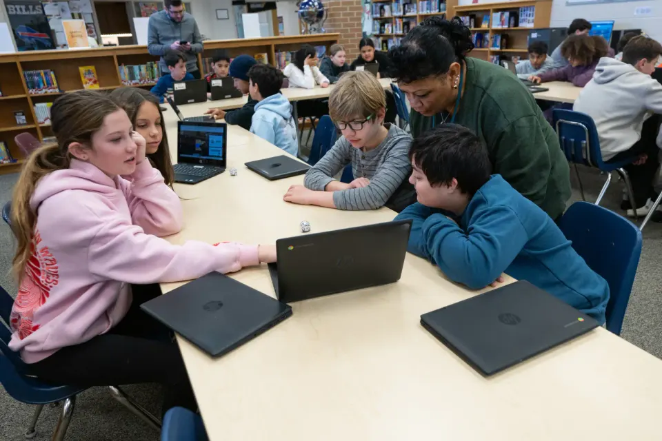 A group of students and a teachers' aide hunch over laptops at a library table.