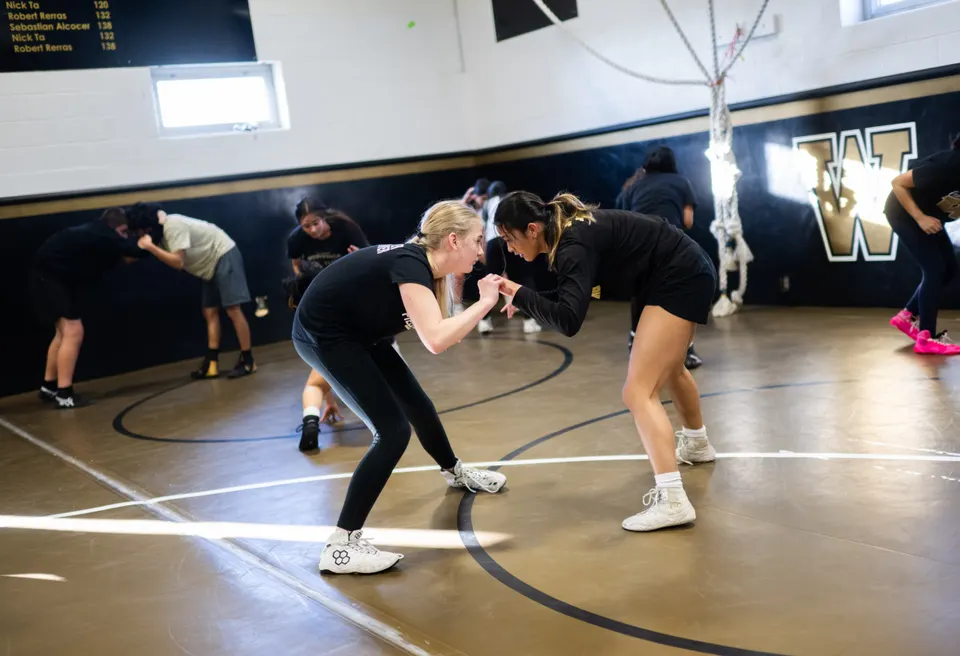 Two girls lock hands, hunched over, making eye contact, on the wrestling mat.